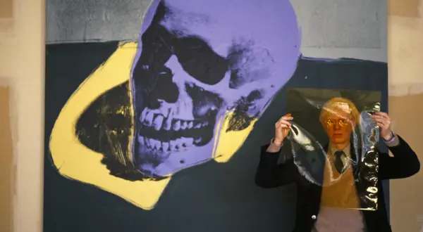 Andy Warhol trzyma przezroczystą folię przed fioletowo-żółtym obrazem czaszki.
